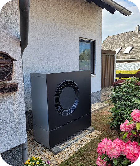 Außenansicht eines weißen Hauses mit einer modernen schwarzen Wärmepumpe auf Kiesbett neben blühenden pinken Blumen.