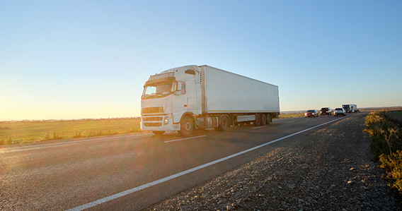 Weißer Lastwagen fährt auf einer Landstraße im Sonnenuntergang mit mehreren Fahrzeugen im Hintergrund.