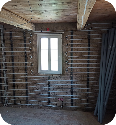 Innenraum mit unverputzter Wand, an der mehrere Heizungsrohre für eine Fußbodenheizung installiert sind, unter einem kleinen Fenster, mit sichtbaren Holzbalken an der Decke.