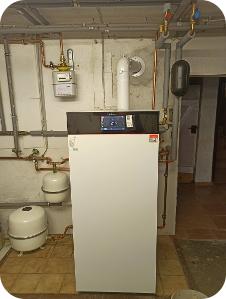 Weiße Heizungsanlage mit digitalem Thermostatdisplay in einem Keller mit an der Wand verlaufenden Rohrleitungen.