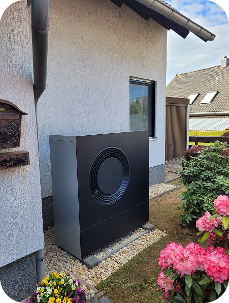 Modern black heat pump unit installed outside a white house, surrounded by gravel and flowering plants.