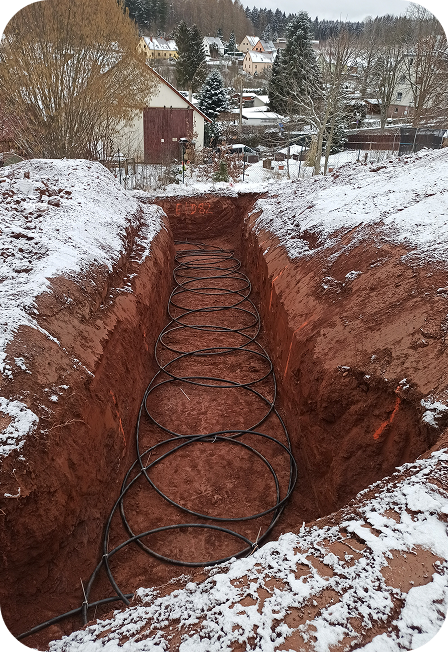 Ein langes, rechteckiges Erdloch mit darin verlegten schwarzen Rohren in einer verschneiten Winterlandschaft mit Häusern und Bäumen im Hintergrund.
