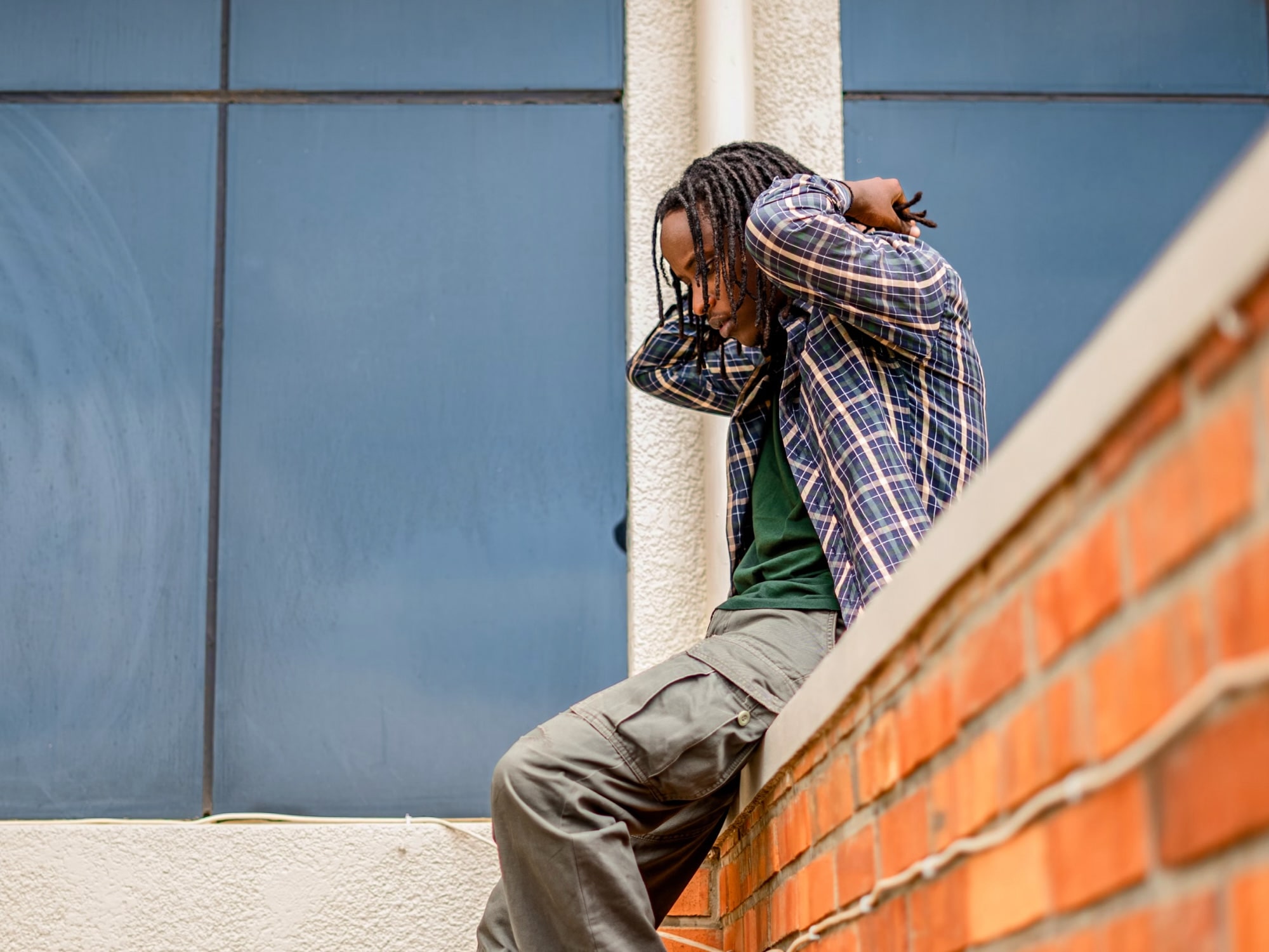 Young person with dreadlocks sitting on a ledge adjusting their hair, wearing a plaid shirt and cargo pants.
