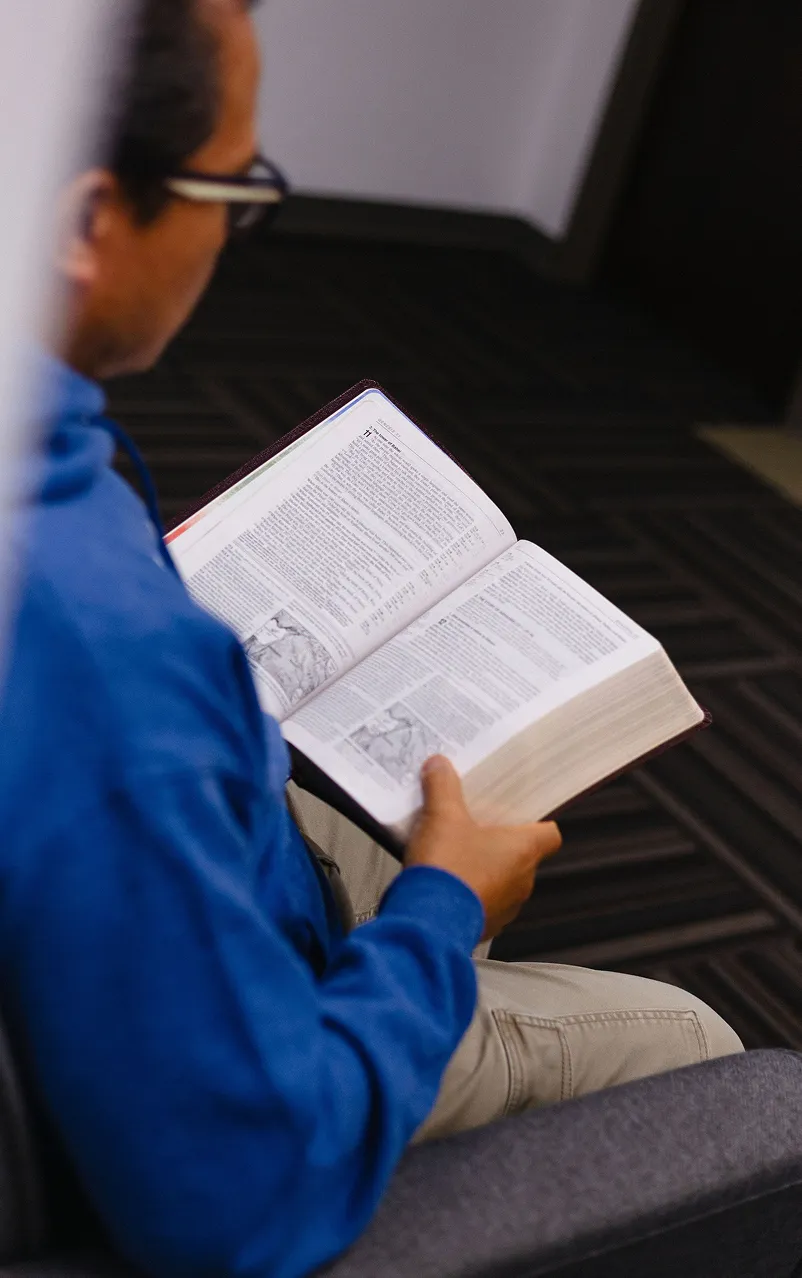 Person wearing glasses and a blue shirt reading an open Bible with text and illustrations.