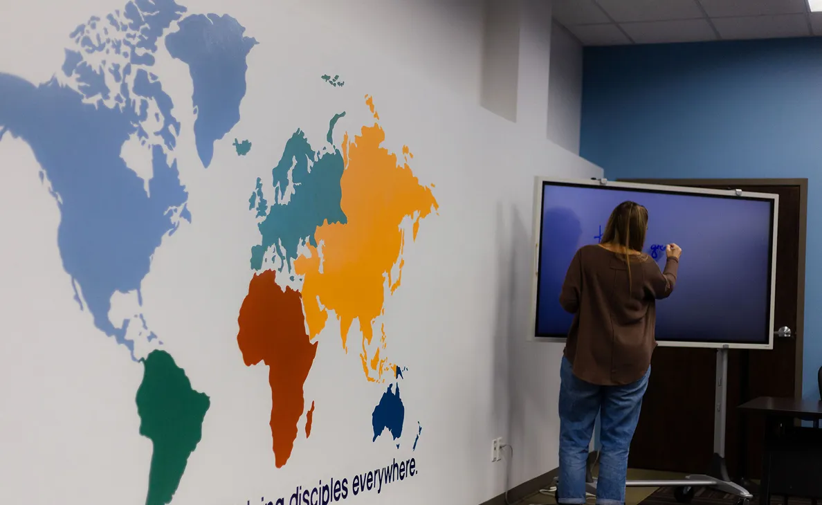 Person writing on a digital touchscreen in a room with a colorful world map mural on the wall.