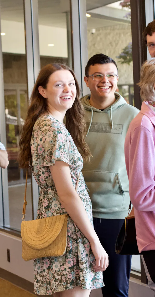 Smiling young woman in a floral dress and small purse standing next to a smiling young man in glasses and a green hoodie indoors.