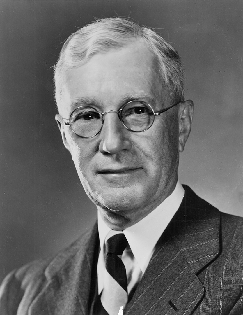 Black and white portrait of older man with round glasses in a suit and striped tie.