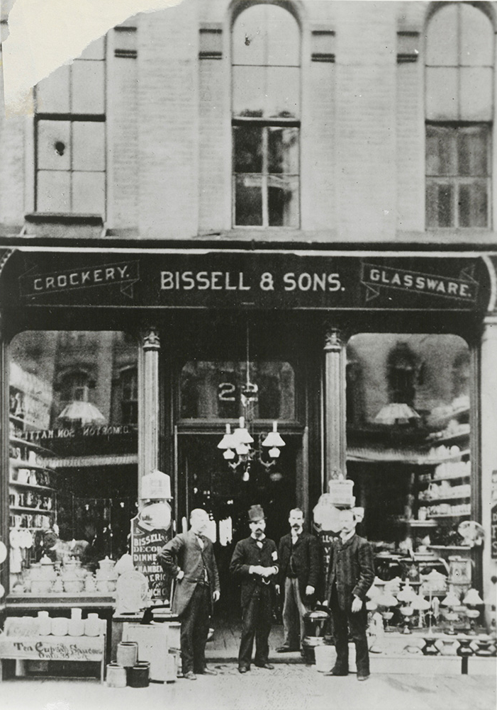 Four men in suits standing outside Bissell & Sons crockery and glassware shop with displayed items.