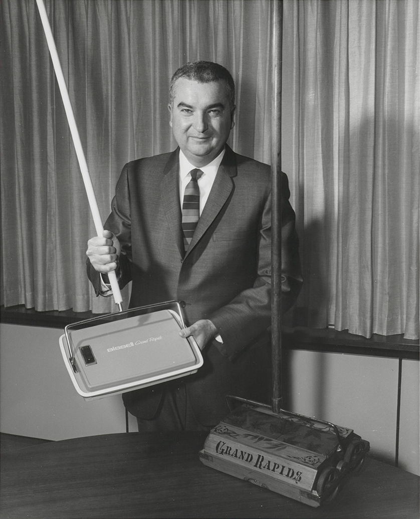 Man in suit holding a Bissell Grand Rapids vacuum cleaner part next to an older vacuum model.