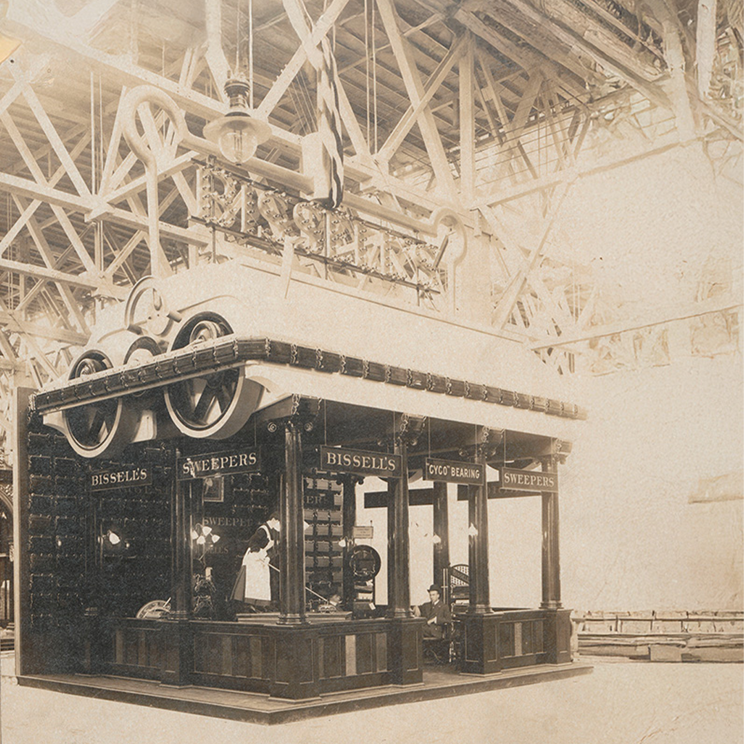 Vintage sepia photo of a Bissell's Sweepers exhibition booth indoors with two people inside.