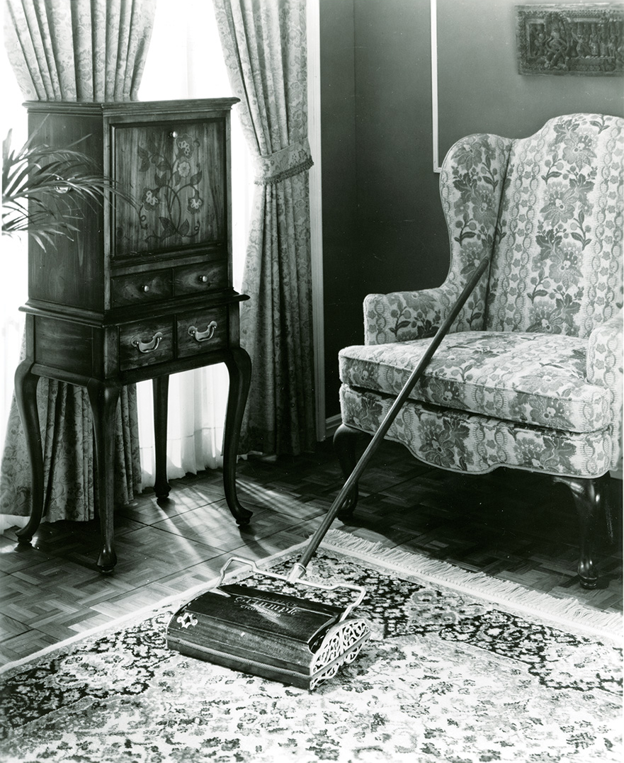 Vintage carpet sweeper with long handle on a patterned rug between floral armchair and wooden cabinet.