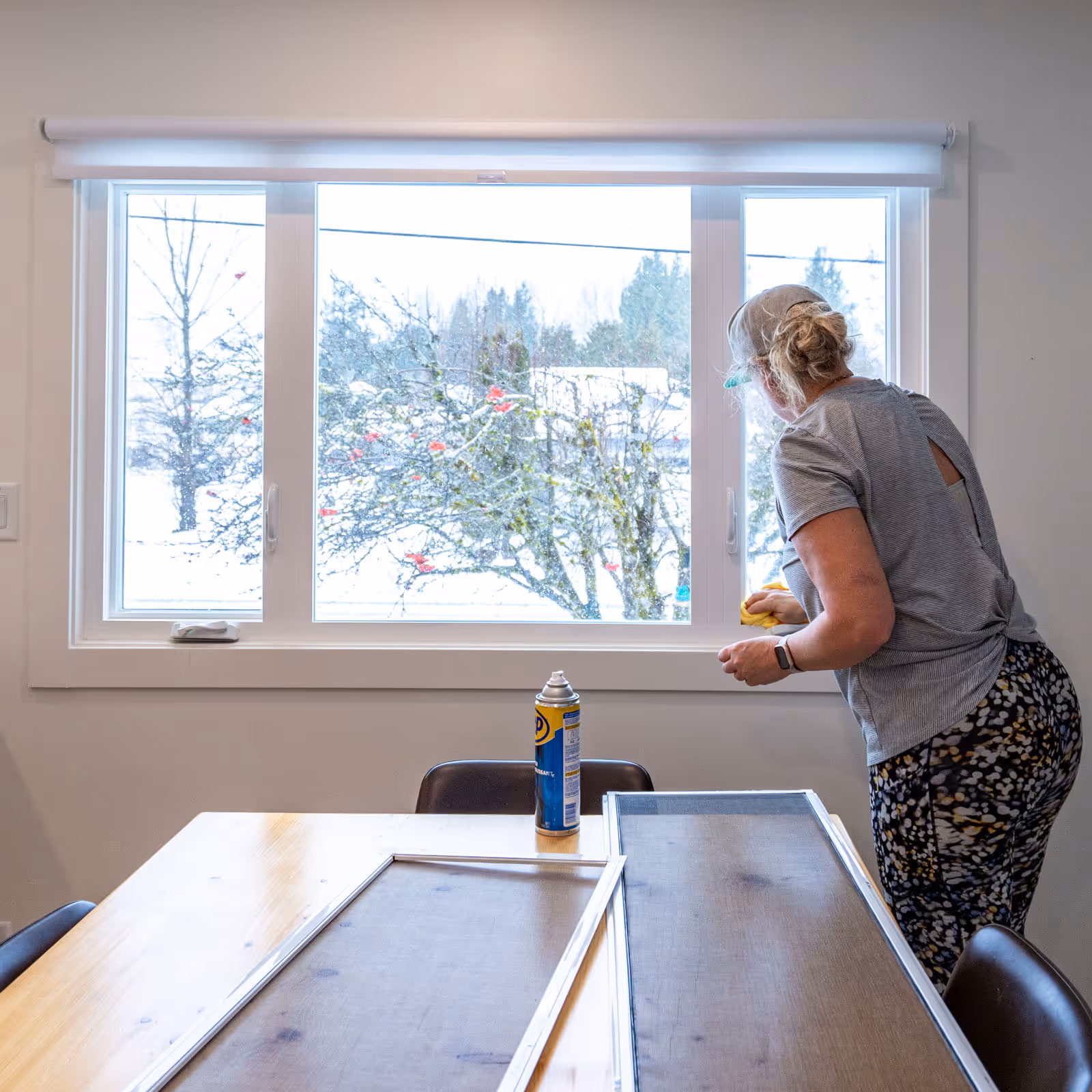 Person cleaning a large window indoors with screens and a can of spray lubricant on the table.