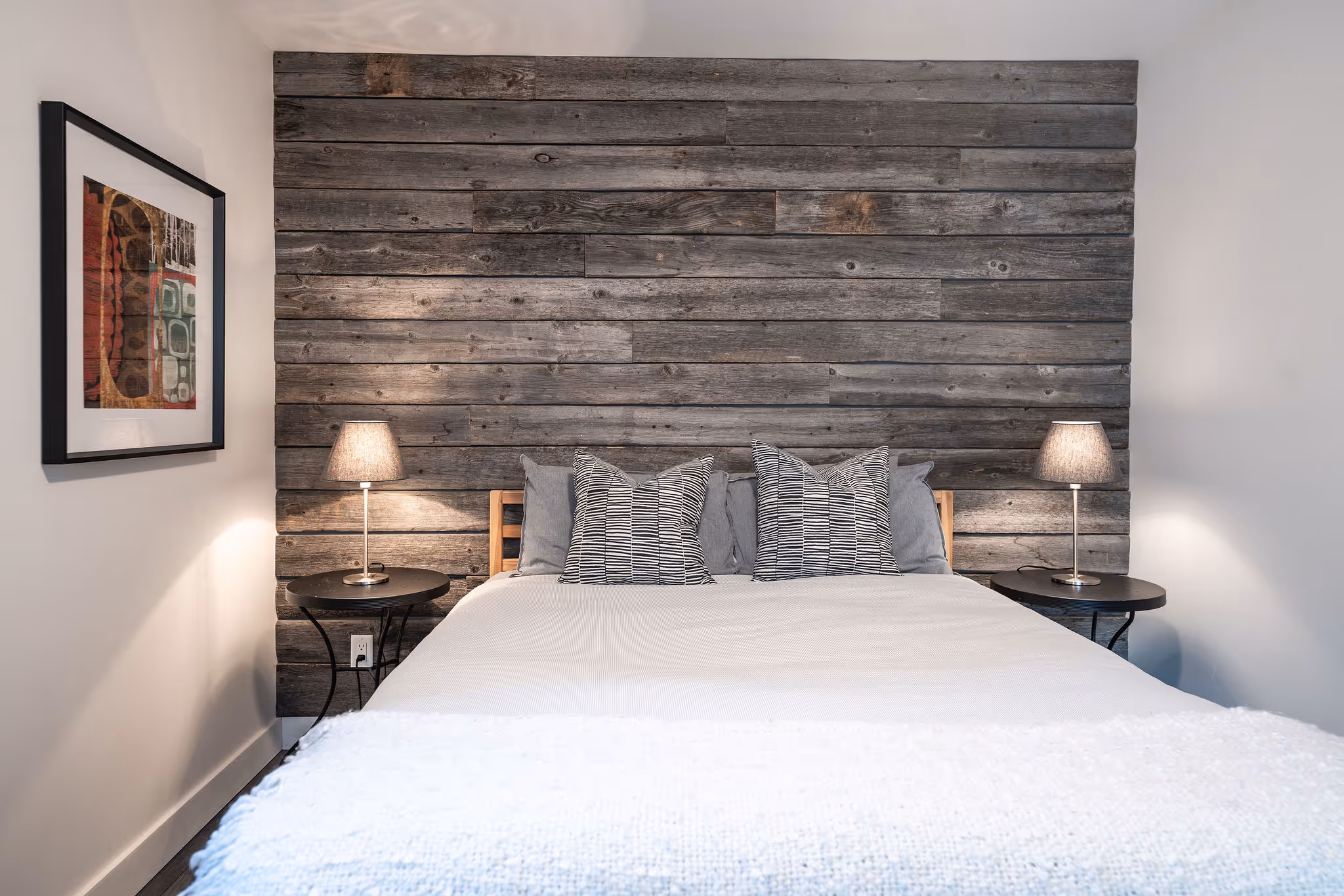 Modern bedroom with a bed featuring white bedding and striped pillows, flanked by two nightstands with table lamps, against a rustic wooden plank accent wall.