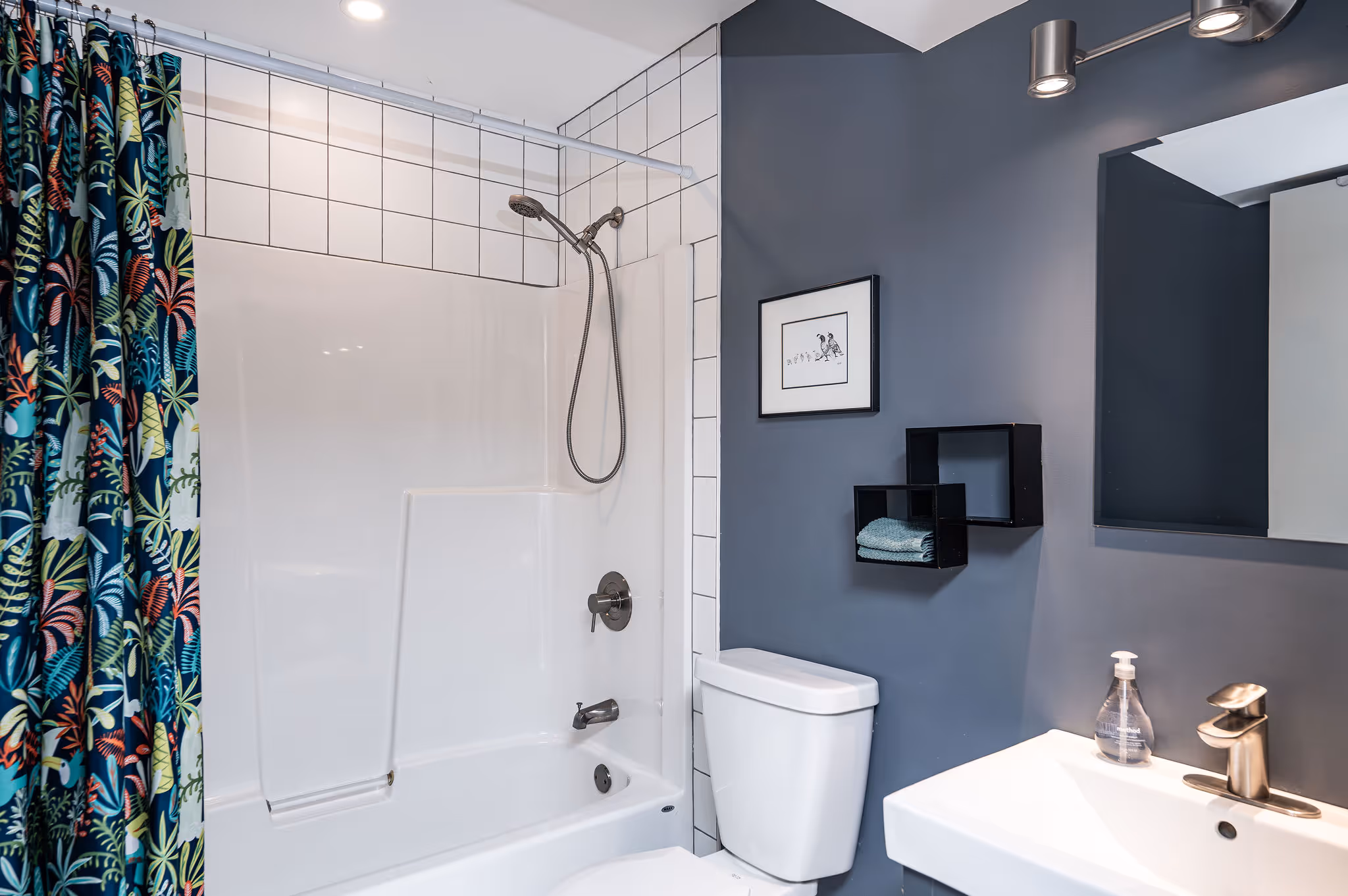 Modern bathroom featuring a white bathtub with tiled walls and a colorful floral shower curtain, a white toilet, a white sink with a soap dispenser, a framed picture, and dark gray walls with black shelves.