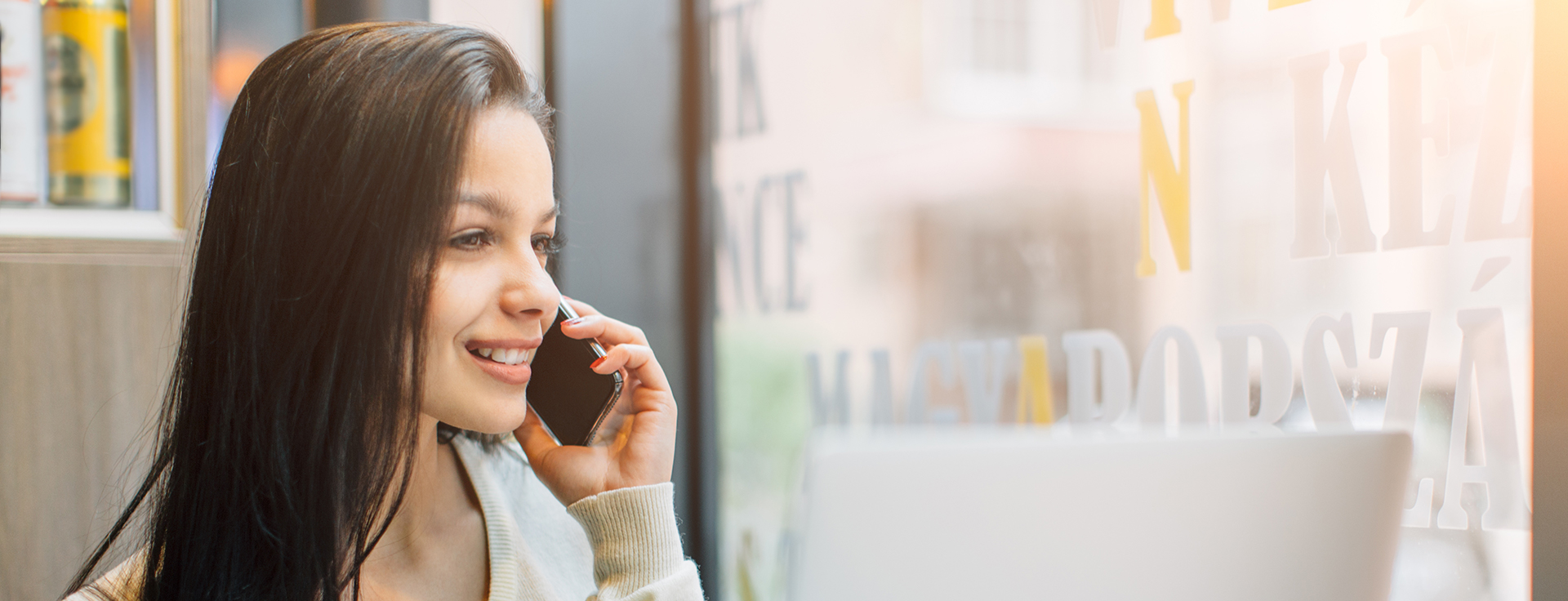 Smiling woman with long dark hair talking on a smartphone near a window with blurred text and a laptop in front of her.
