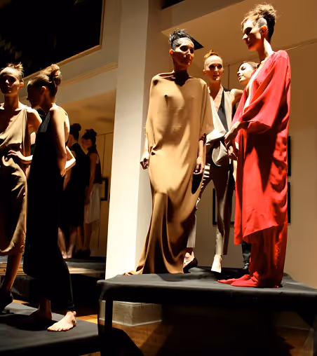 Several fashion models wearing minimalist dresses and outfits standing on elevated platforms in a dimly lit setting.