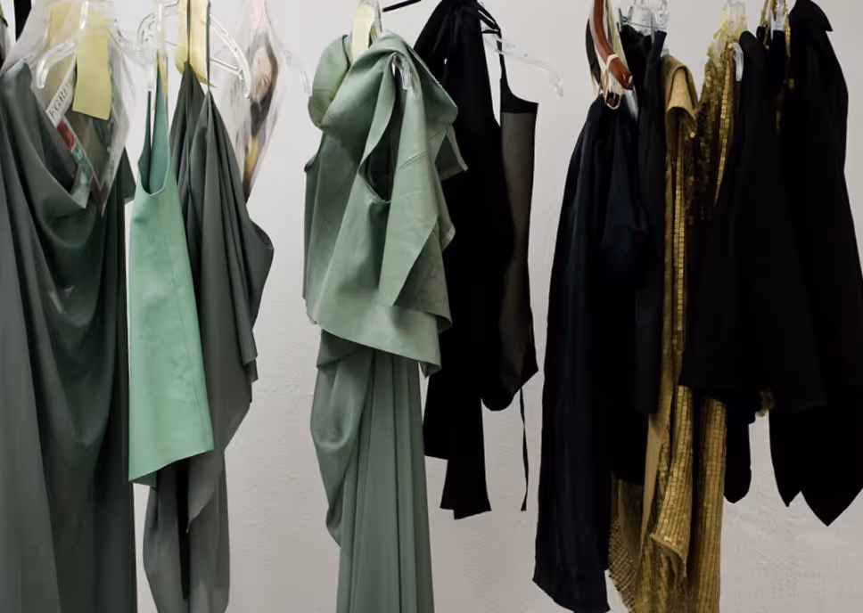 Various dresses in green, black, and gold hanging on clear plastic hangers against a white wall.