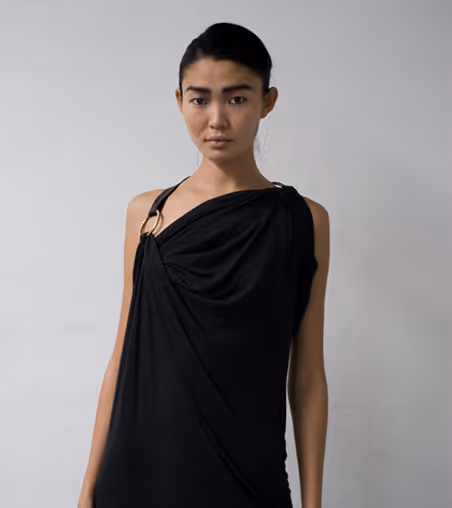 Woman with dark hair pulled back wearing a black draped dress with a ring detail on the shoulder against a plain gray background.