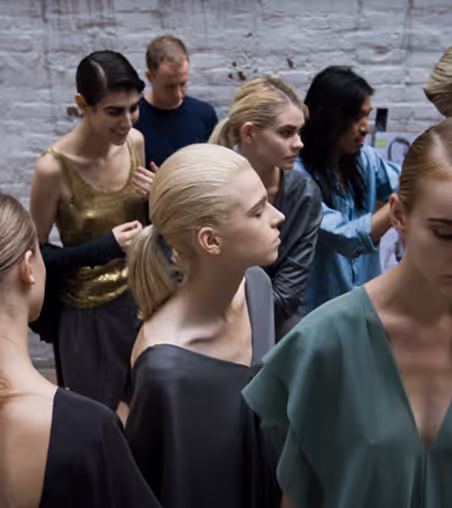 Group of young models with pulled-back hair wearing varied modern outfits standing and interacting indoors against a white brick wall.