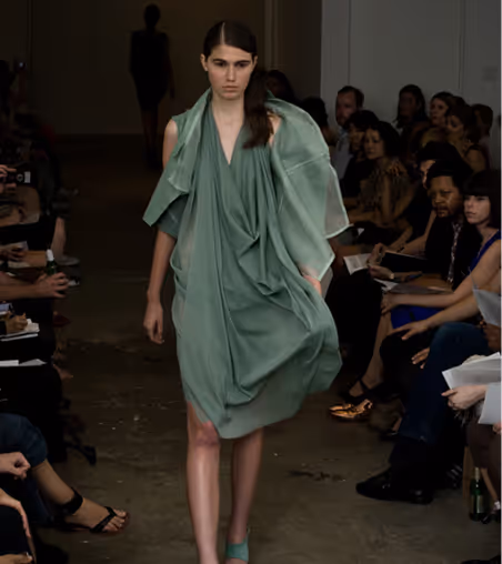 Model walking on runway wearing a draped sage green dress during a fashion show.