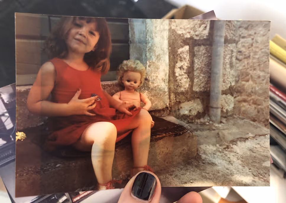 A photograph held by a hand shows a young girl in a red dress sitting on stone steps next to a doll with blond curly hair.