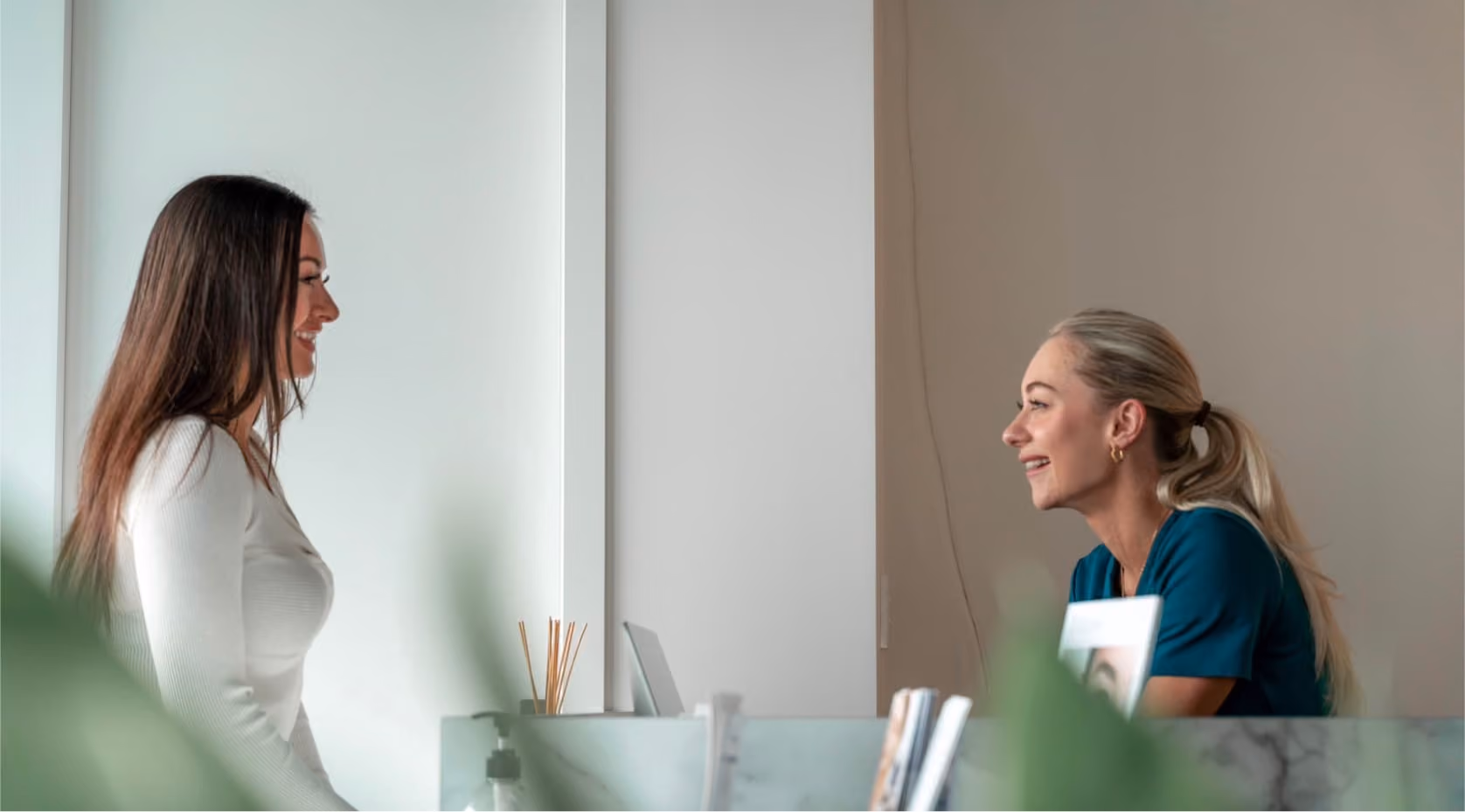 Two women smiling and talking across a reception desk in a bright, modern office.