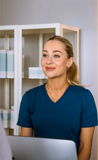 Smiling woman with blonde hair in a ponytail wearing teal medical scrubs sitting at a desk with a laptop in a clinical setting.