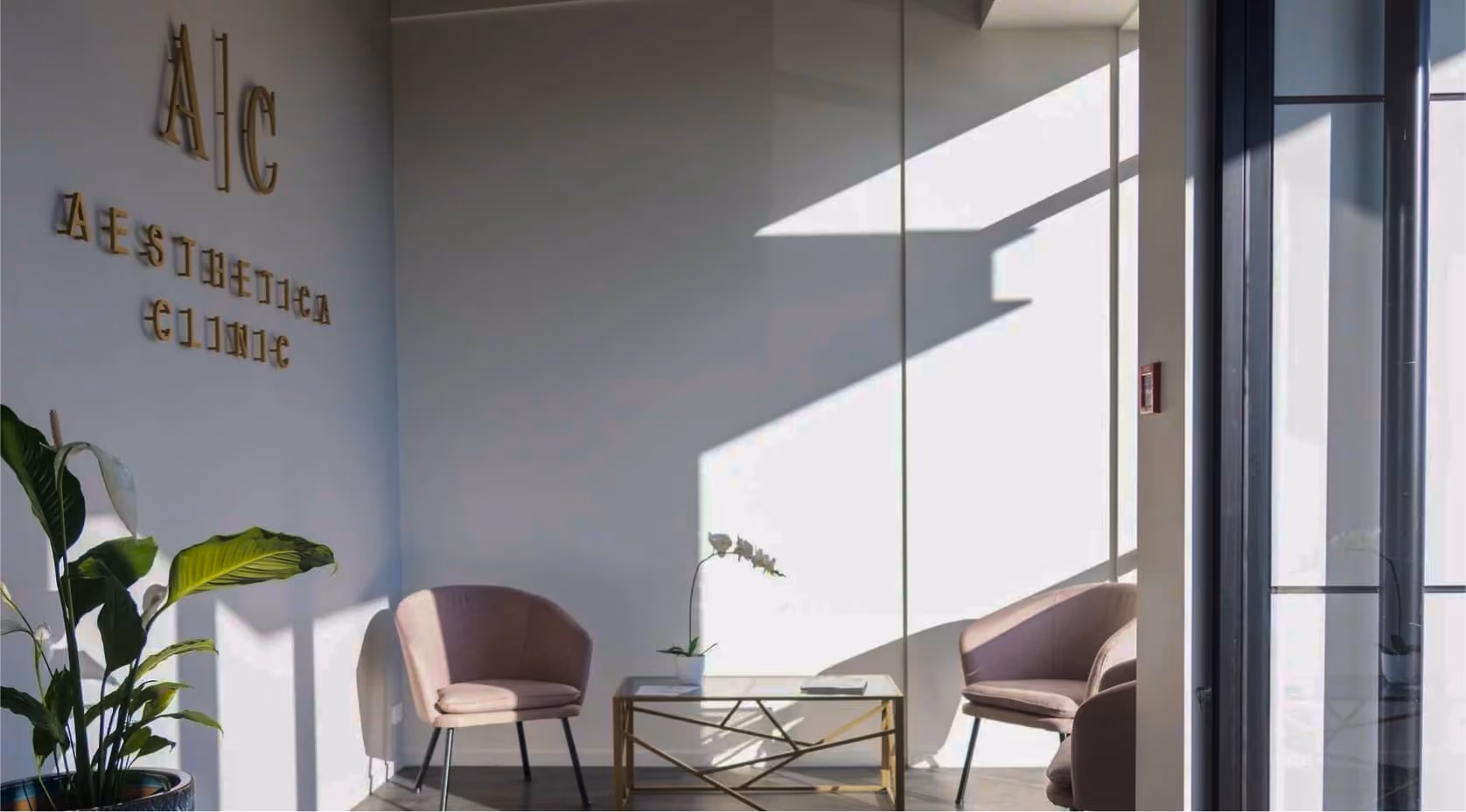 Sunlit waiting area with two pink chairs, a glass-top table with a small plant, and a wall sign reading 'A1C Aesthetica Clinic'.