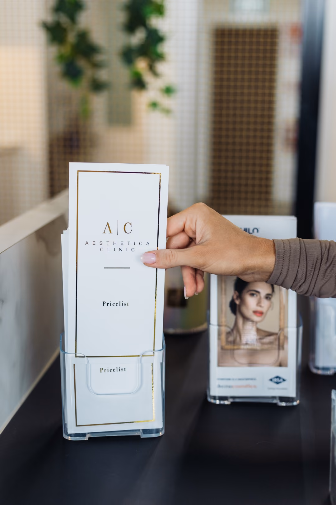 A hand with manicured nails holding a white brochure labeled 'Aesthetica Clinic Pricelist' in a clear stand, with another brochure holder in the background featuring a woman's face.