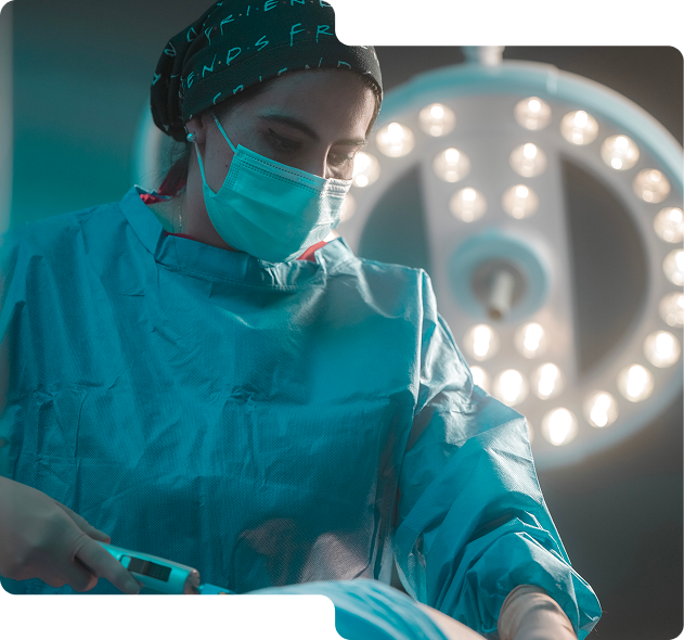 Médica con gorro y mascarilla quirúrgica realizando una cirugía bajo luces brillantes en un quirófano.