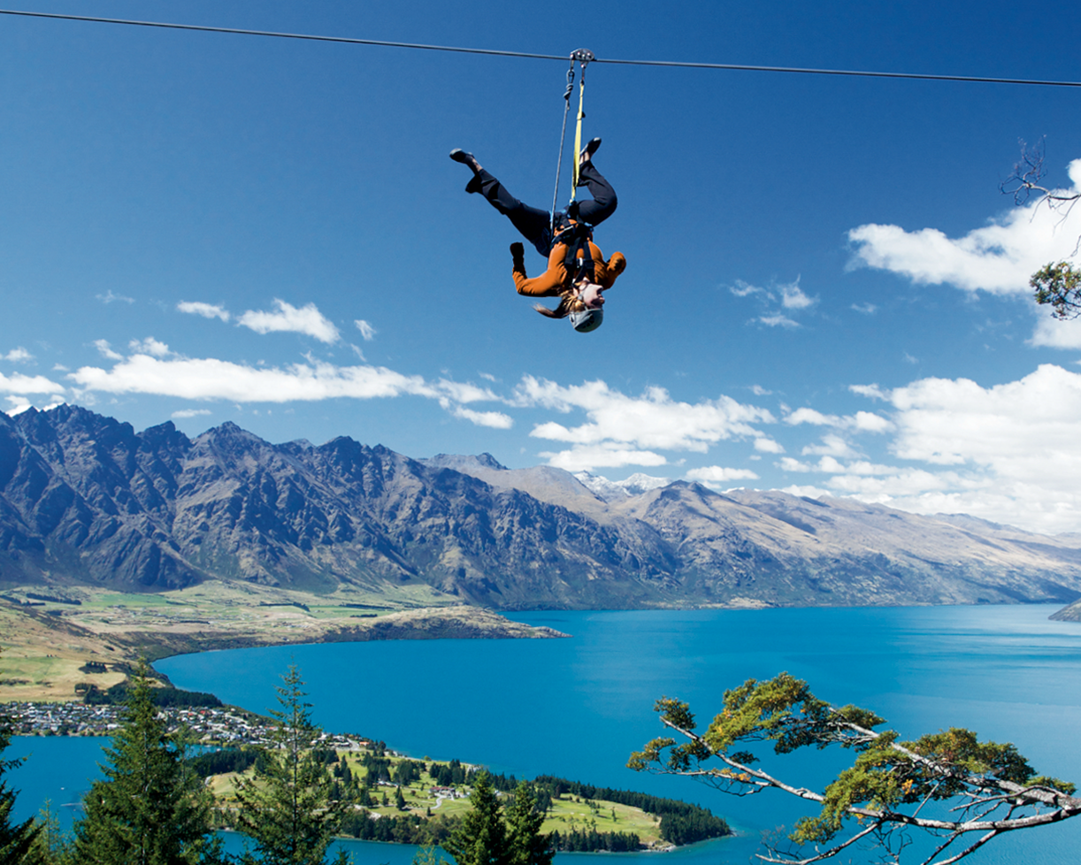 Person riding upside down on a zip line in an action pose with mountains in the background