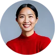 Smiling Asian woman with long black hair wearing a red turtleneck sweater against a gray background.