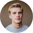 Headshot of a young man with short blond hair wearing a light gray shirt against a blurred brown background.