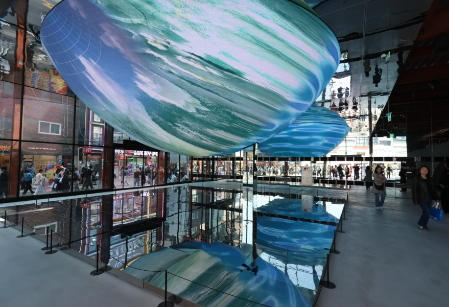 Visitors at Blue Elephant Space Seongsu standing beneath the 12-meter LED sphere displaying flowing blue media art, reflected on the mirrored floor