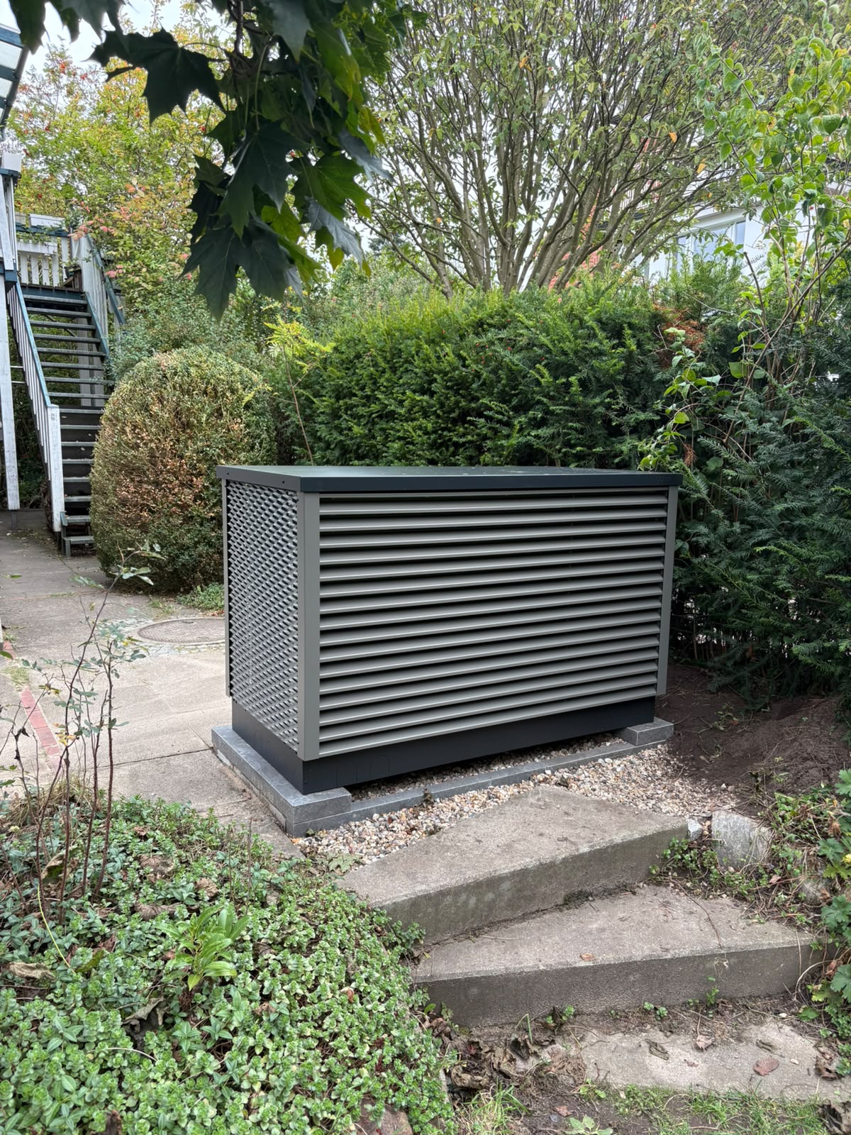 OVUM Wärmepumpe Außeneinheit mit Lamellenverkleidung auf Fundament im Kiesbett in einem Garten in Hamburg
