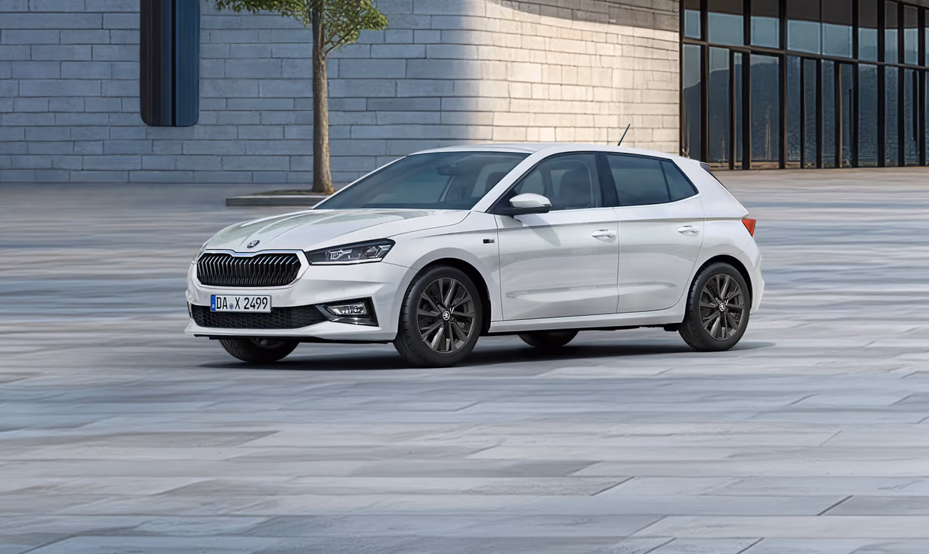 Seitenansicht eines Škoda Fabia Balance in der Lackierung Moon-Weiß Perleffekt vor einer modernen, grauen Gebäudefassade. Das Fahrzeug ist mit dunklen 16-Zoll-Leichtmetallfelgen Proxima (schwarz glanzgedreht) ausgestattet. Auf dem Bild ist ein grüner Störer platziert, der einen Preisvorteil in Höhe von 1.800€ sowie 5 Jahre Garantieleistung bewirbt. 