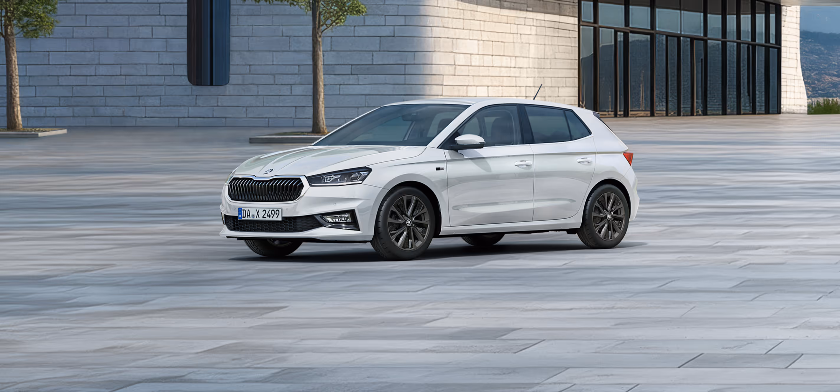 Seitenansicht eines Škoda Fabia Balance in der Lackierung Moon-Weiß Perleffekt vor einer modernen, grauen Gebäudefassade. Das Fahrzeug ist mit dunklen 16-Zoll-Leichtmetallfelgen Proxima (schwarz glanzgedreht) ausgestattet. Auf dem Bild ist ein grüner Störer platziert, der einen Preisvorteil in Höhe von 1.800€ sowie 5 Jahre Garantieleistung bewirbt. 