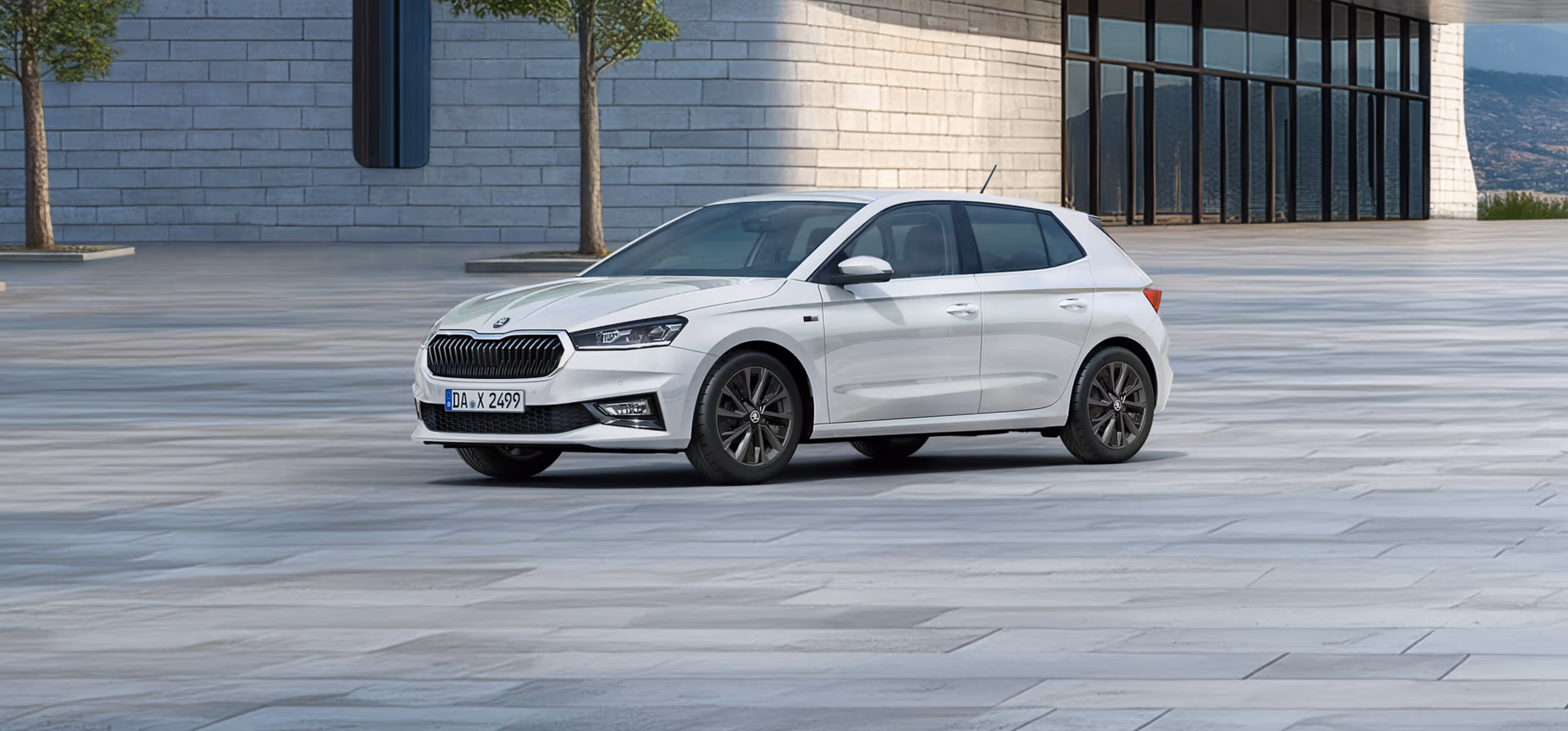 Seitenansicht eines Škoda Fabia Balance in der Lackierung Moon-Weiß Perleffekt vor einer modernen, grauen Gebäudefassade. Das Fahrzeug ist mit dunklen 16-Zoll-Leichtmetallfelgen Proxima (schwarz glanzgedreht) ausgestattet. Auf dem Bild ist ein grüner Störer platziert, der einen Preisvorteil in Höhe von 1.800€ sowie 5 Jahre Garantieleistung bewirbt. 