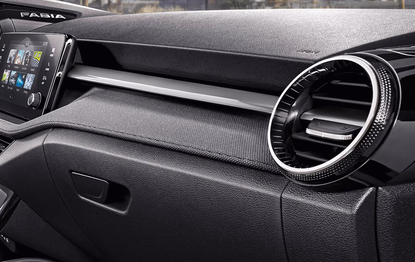 Detailaufnahme des Armaturenbretts auf der Beifahrerseite des Škoda Fabia Balance. Das Bild zeigt die runde, chromverzierte Lüftungsdüse sowie die Dekorleiste mit grauem Stoffbezug. Darunter befindet sich das Handschuhfach und links ist der Rand des Infotainment-Displays angeschnitten.