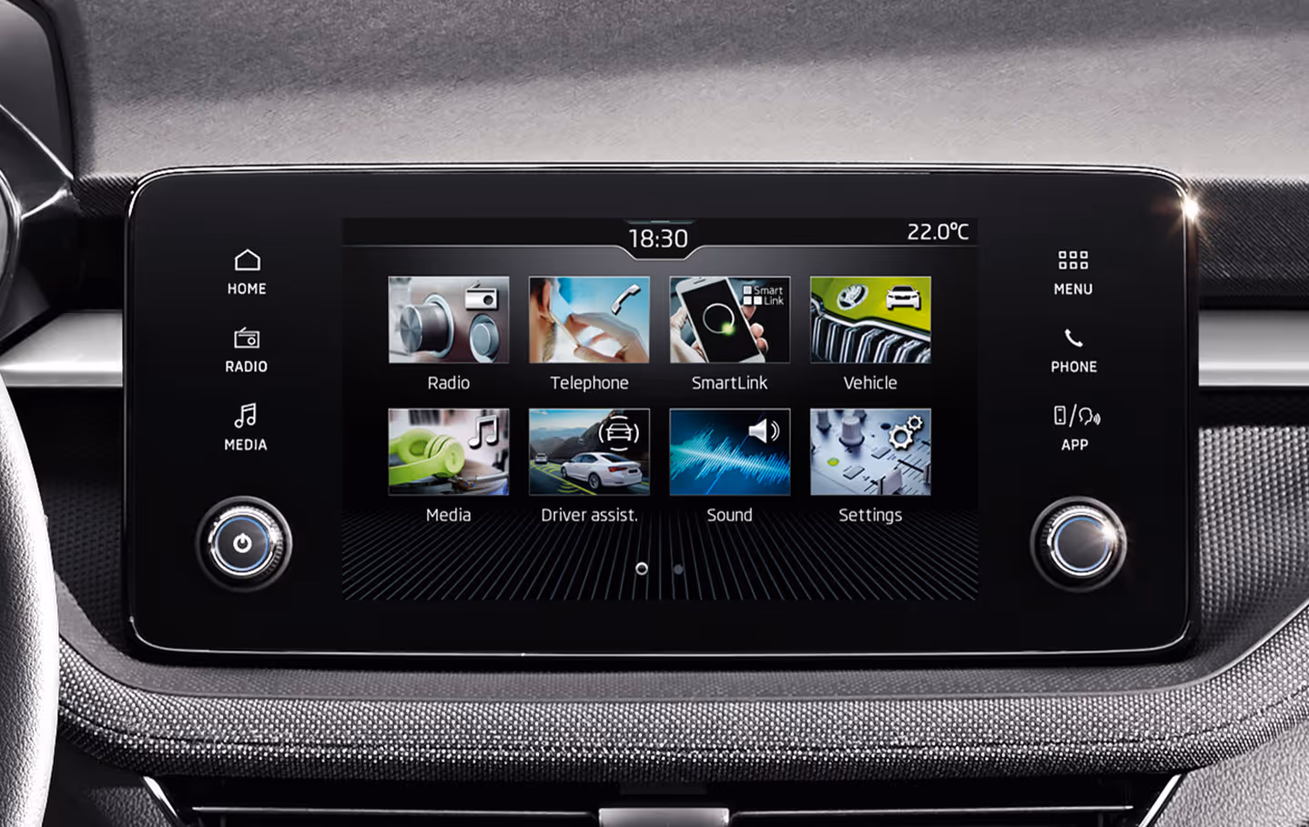 Nahaufnahme des zentralen Infotainment-Displays im Škoda Fabia Balance, das im Hauptmenü verschiedene Funktionskacheln wie Radio, Telefon und SmartLink anzeigt. Der Touchscreen wird seitlich von Sensortasten und zwei physischen Drehreglern flankiert. Unterhalb der Einheit ist die Dekorleiste mit dem grauen Stoffbezug sichtbar.