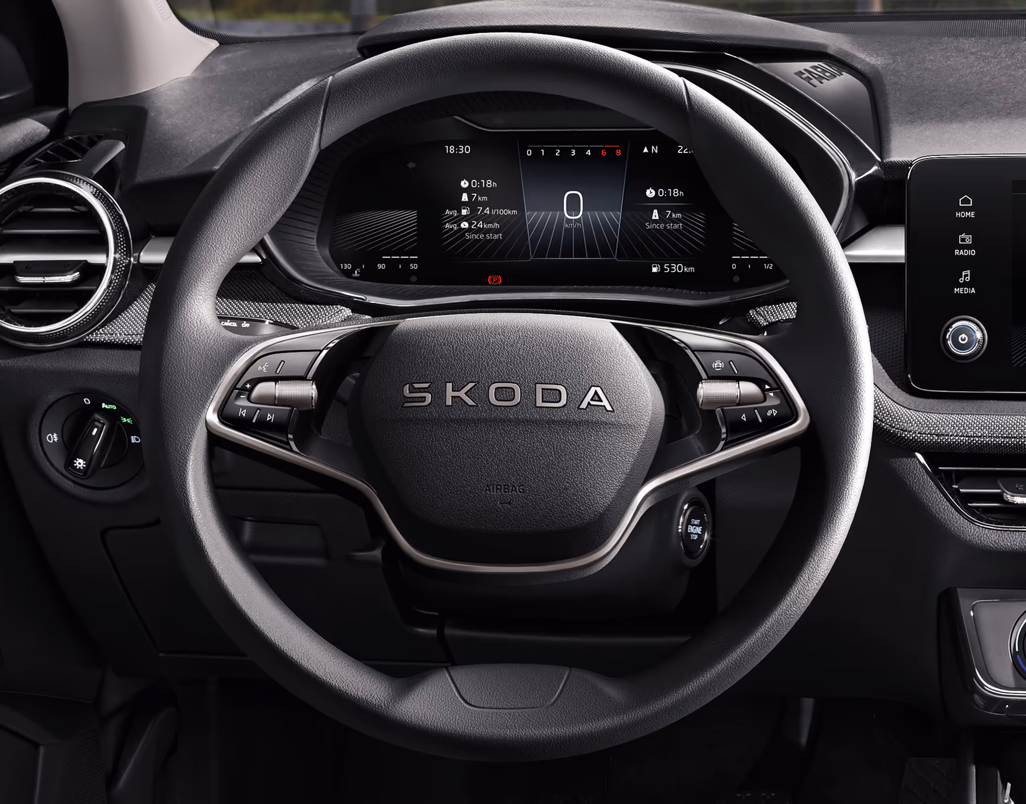 Nahaufnahme des 2-Speichen-Multifunktionslenkrads im Škoda Fabia Balance mit silbernen Bedienelementen und zentralem Škoda Schriftzug. Der Blick fällt durch das Lenkrad auf das digitale Kombiinstrument, das aktuelle Fahrdaten anzeigt. Links daneben ist die runde, chromverzierte Lüftungsdüse in das Armaturenbrett eingelassen.