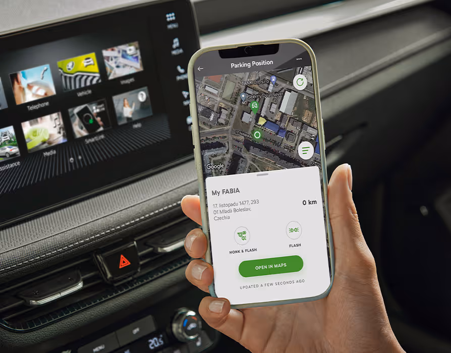 Nahaufnahme einer Hand, die ein Smartphone mit der geöffneten MyŠkoda App hält, welche den aktuellen Parkstandort („Parking Position“) und Fahrzeugstatus anzeigt. Im Hintergrund ist das Armaturenbrett mit dem Infotainment-Display des Škoda Fabia Balance unscharf erkennbar.