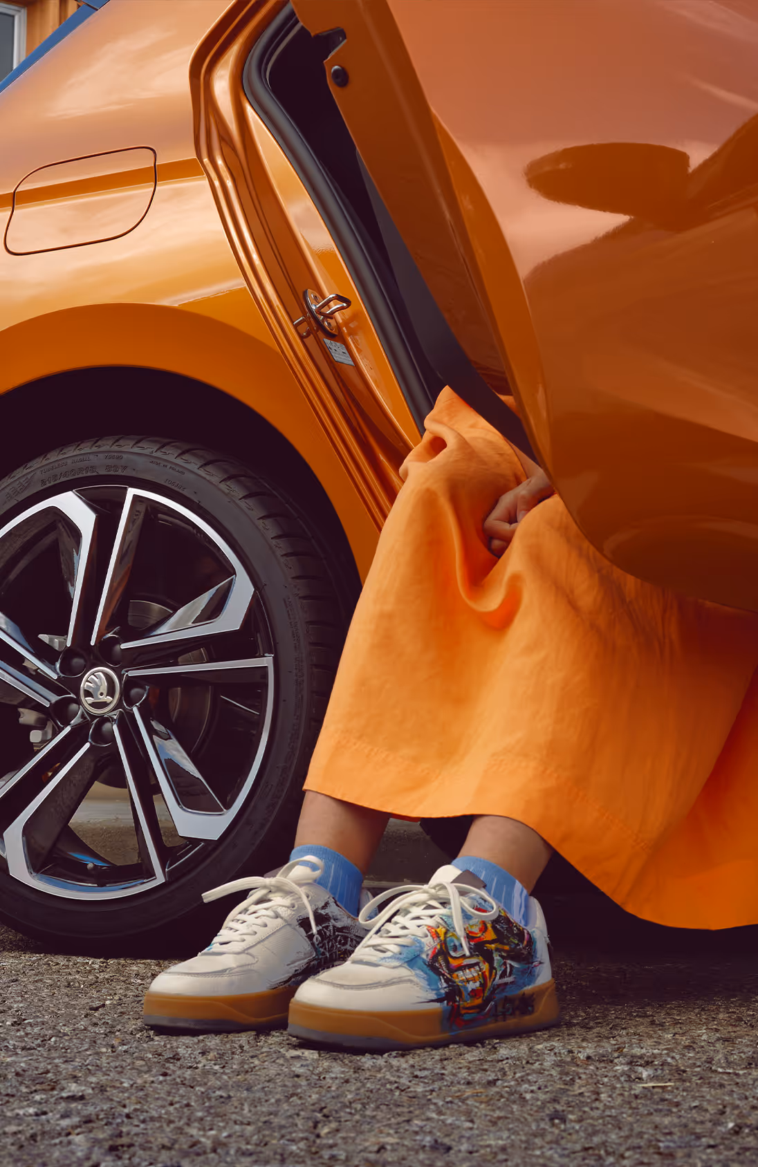 Eine Detailaufnahme zeigt den hinteren Bereich eines Škoda Fabia in der Lackierung Phoenix-Orange Metallic, aus dessen geöffneter Fondtür eine Person in einem farblich abgestimmten Rock steigt. Im Fokus steht dabei die glanzgedrehte Leichtmetallfelge mit ihren markanten Aero-Einsätzen. Modische Sneaker und eine urbane Gebäudefassade im Hintergrund runden die Szenerie ab.