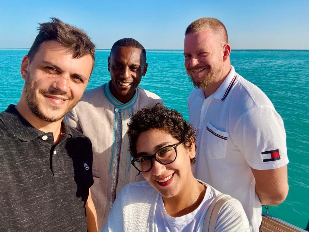 Čtyři lidé usmívající se na kameru se stojí u modrého moře za slunečného dne.