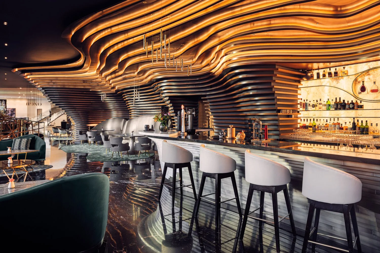 Modern bar interior with wavy illuminated ceiling design, white bar stools, and shelves stocked with liquor bottles.