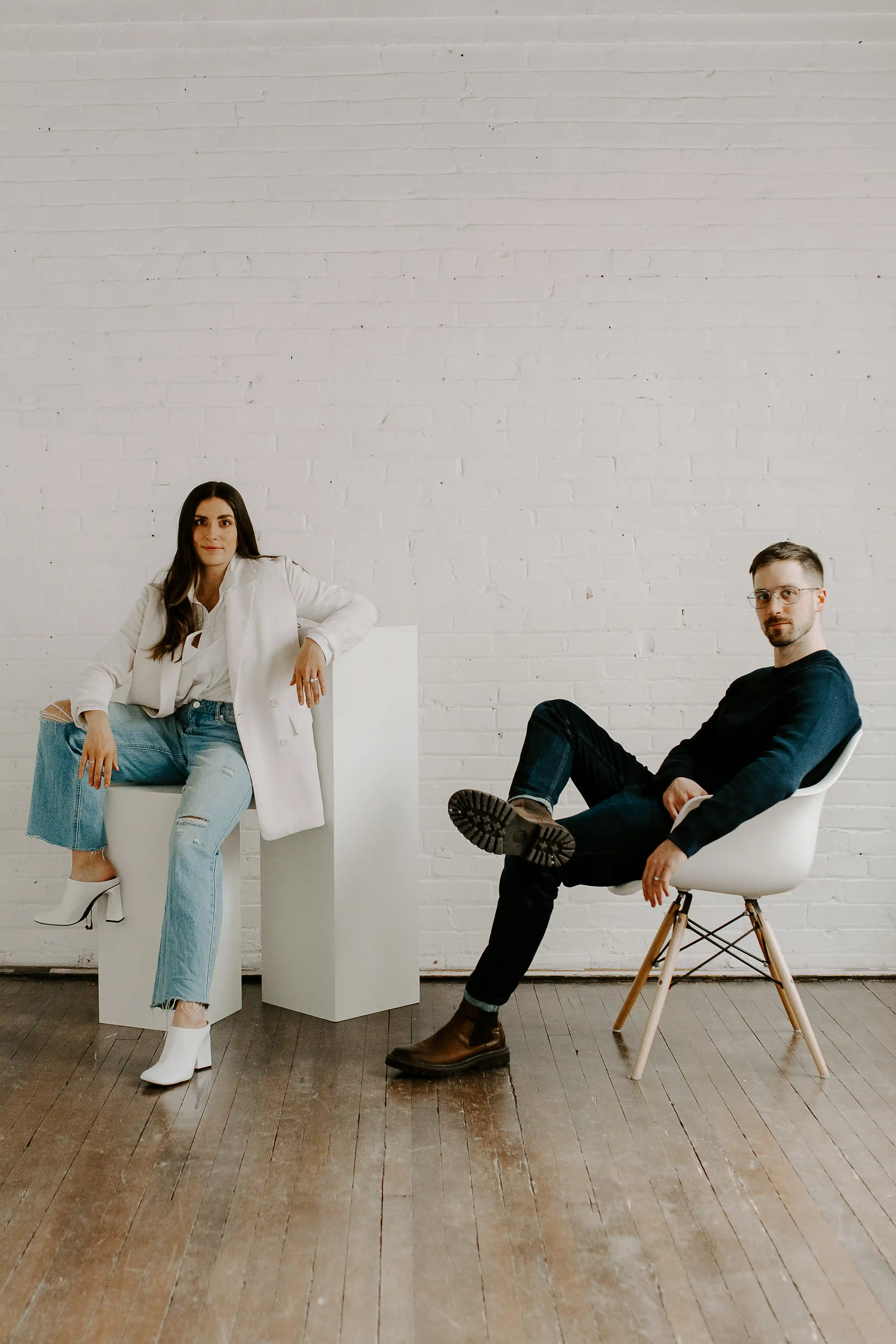 Portrait of the ofLight Design founders, Justine Schmode and Bryce Schmode, posing in a bright, modern studio with white painted brick walls and hardwood floors. The woman sits on a minimalist white architectural plinth wearing a white blazer and denim, while the man sits in a classic mid-century molded chair.