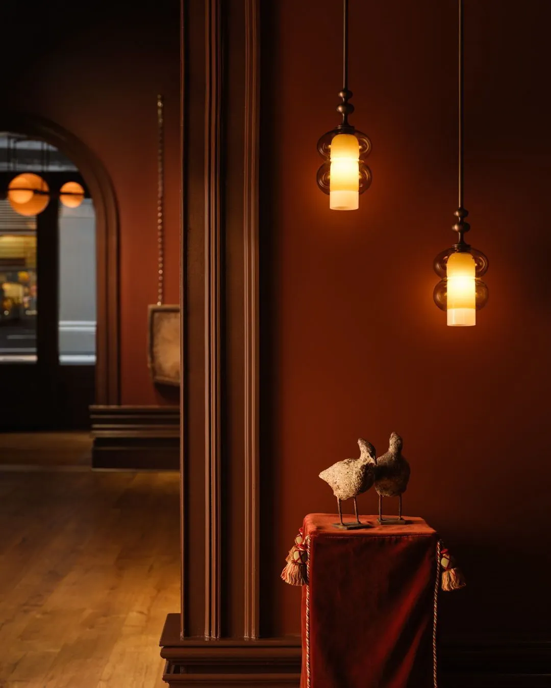 SkLO + AvroKO Prince pendant lights, featuring layered amber and frosted glass cylinders, suspended in a moody terracotta interior above a red velvet display pedestal.