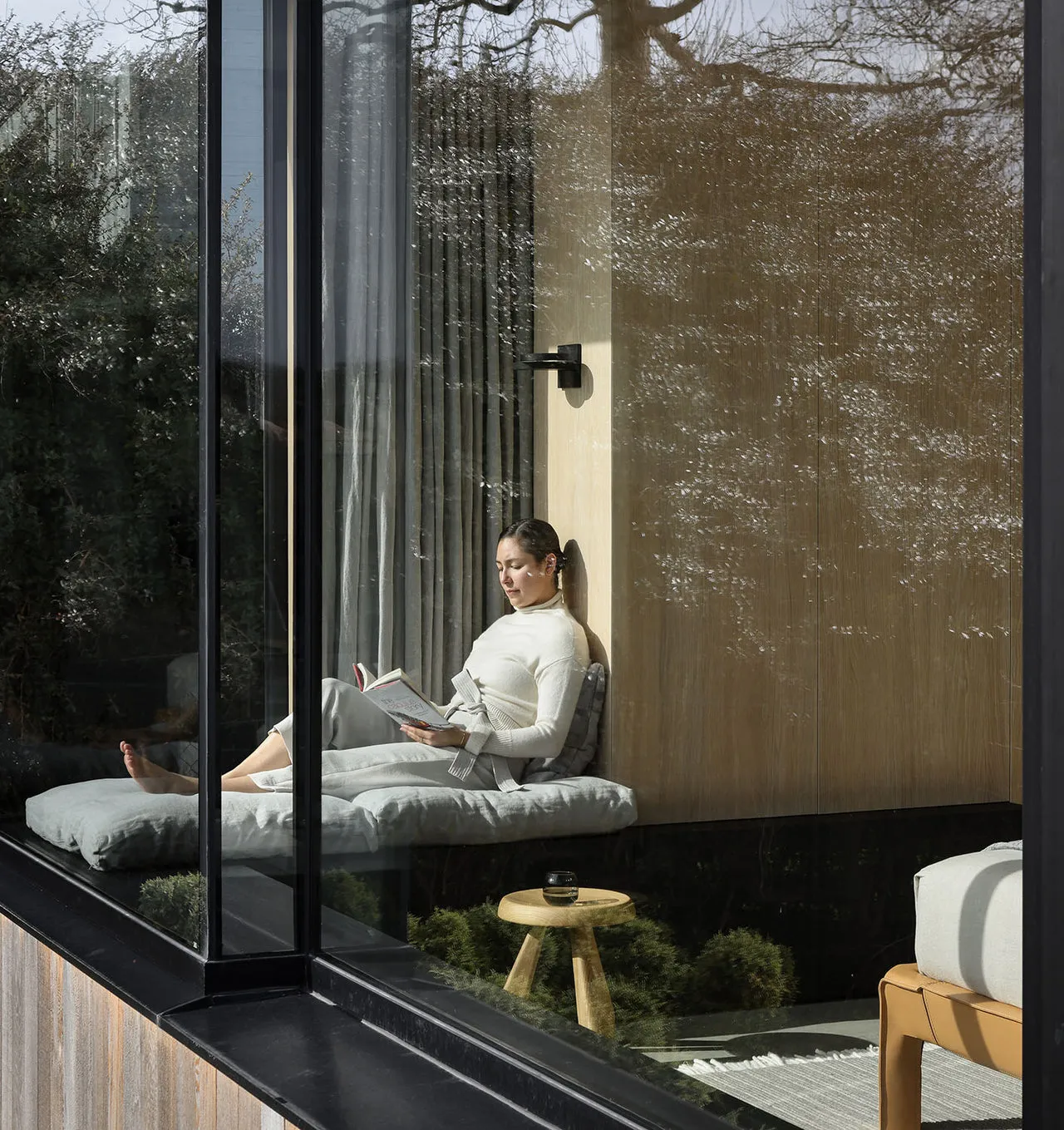 Tomnuk Lune Wall Sconce, a sleek black minimalist wall light mounted on light wood paneling above a cozy, sunlit window seat where a woman is reading, viewed from the outside through large architectural glass windows reflecting bare trees.