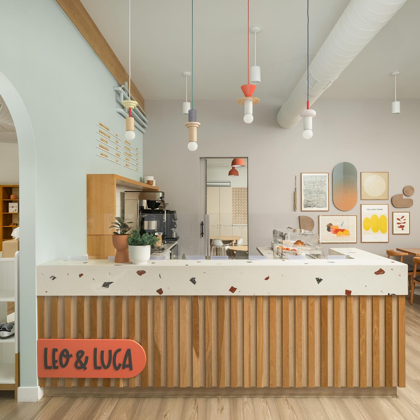 Schneid Studio Junit pendant lights, a trio of playful, geometric wooden suspension fixtures with colorful painted elements and exposed bulbs, hanging above a terrazzo coffee bar with a ribbed wood base in a bright, modern cafe interior.