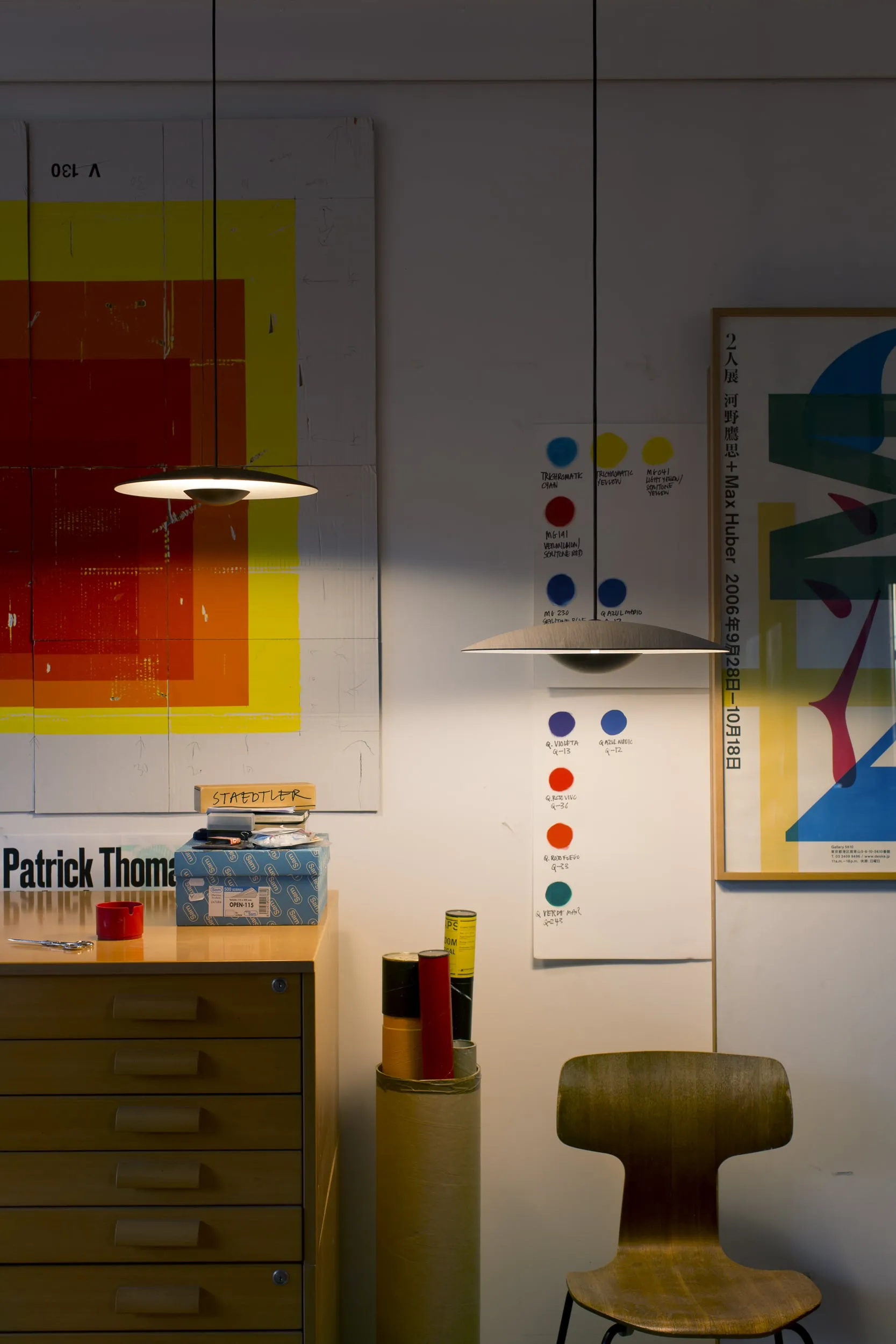 Here is the SEO-optimized alt text for this creative, eclectic shot:

"Marset Ginger pendant lights, a pair of ultra-thin, flat-profile suspension fixtures casting warm downlight in an eclectic art studio workspace surrounded by colorful abstract posters, a wooden flat-file cabinet, and a mid-century plywood chair.